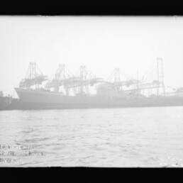 Frachtschiff am Kai mit mehreren großen Hafenkränen im Dunst, Wasser im Vordergrund; Museumslogo unten links.