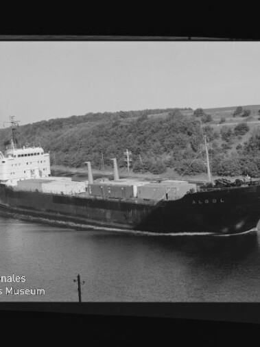 Frachtschiff mit Namenszug am Bug auf einem Kanal vor bewaldetem Ufer, mit Wasserzeichen des Int. Maritimen Museums Hamburg
