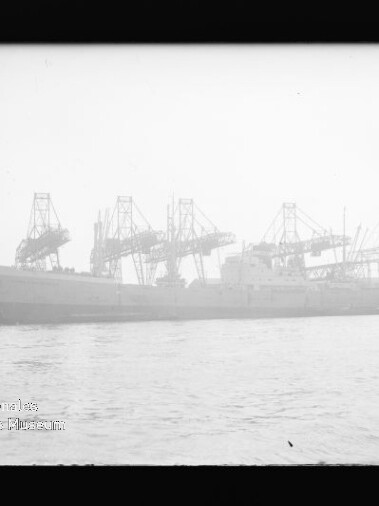 Großes Frachtschiff im Hafen vor mehreren hohen Hafenkränen, Wasser im Vordergrund und Museumswasserzeichen links unten.