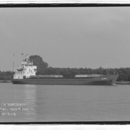 Schwarzweißfoto eines Frachtschiffs CLARA auf ruhigem Wasser vor Baumreihe, mit Wasserzeichen Maritimes Museum Hamburg