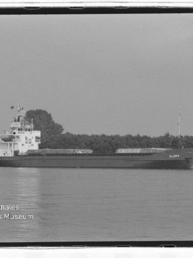 Schwarzweißfoto eines Frachtschiffs CLARA auf ruhigem Wasser vor Baumreihe, mit Wasserzeichen Maritimes Museum Hamburg