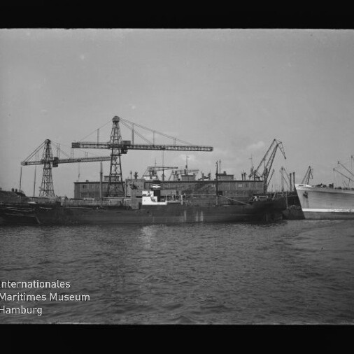Frachtschiff im Hafen vor Werfthallen mit mehreren großen Hafenkränen, Museum-Logo und Schriftzug unten links.