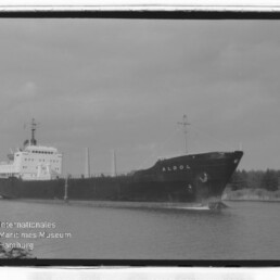 Frachtschiff Algol mit dunklem Rumpf und hellem Aufbau fährt auf ruhigem Kanal, Baumreihe am Ufer; historische Schwarzweißaufnahme.
