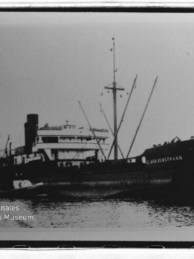 Schwarzweiß-Foto eines Frachtschiffs im Wasser mit Aufbauten, Schornstein, Ladekränen und Spiegelung, Museumslogo unten links.