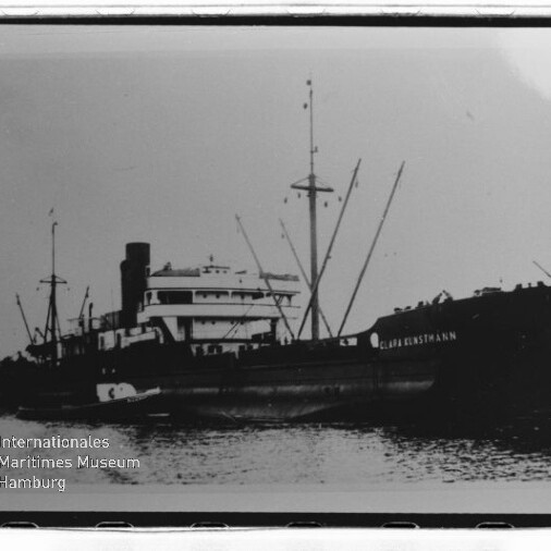 Schwarzweiß-Foto eines Frachtschiffs im Wasser mit Aufbauten, Schornstein, Ladekränen und Spiegelung, Museumslogo unten links.