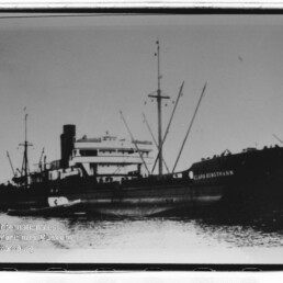 Schwarzweißfoto eines großen Frachtschiffs mit Masten und Aufbauten, ein kleines Beiboot liegt am Rumpf im ruhigen Hafenwasser.