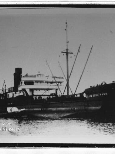 Schwarzweißfoto eines großen Frachtschiffs mit Masten und Aufbauten, ein kleines Beiboot liegt am Rumpf im ruhigen Hafenwasser.