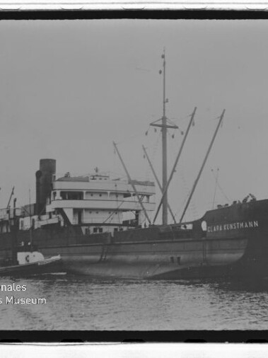 Schwarzweißfoto eines Frachtschiffs mit Schornstein und mehreren Masten, einem Beiboot am Rumpf und Museumslogo links unten.