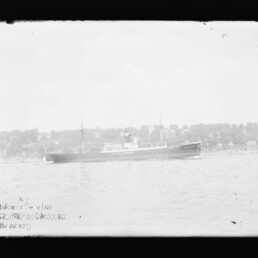 Langes Passagier-/Frachtschiff auf ruhigem Wasser vor einer von Bäumen gesäumten Küste, verblasstes Schwarzweißbild mit dunklem Rahmen.
