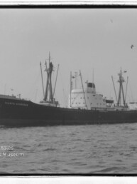 Schwarzweißaufnahme eines Frachtschiffs mit Masten und Schornstein, rechts ein Schlepper, Hafen im Hintergrund.