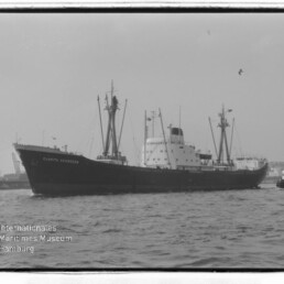 Schwarzweißaufnahme eines Frachtschiffs mit Masten und Schornstein, rechts ein Schlepper, Hafen im Hintergrund.