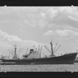 Frachtschiff mit vielen Ladebäumen und mittschiffs liegenden Aufbauten auf ruhiger See unter bewölktem Himmel; Wasserzeichen Internationales Maritimes Museum Hamburg