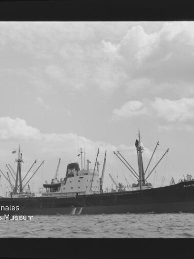 Frachtschiff mit vielen Ladebäumen und mittschiffs liegenden Aufbauten auf ruhiger See unter bewölktem Himmel; Wasserzeichen Internationales Maritimes Museum Hamburg
