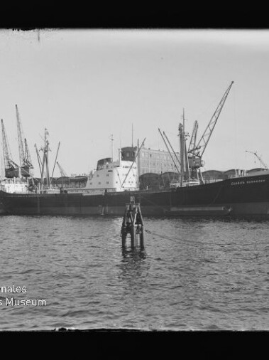 Frachtschiff am Kai mit mehreren Hafenkränen im Hintergrund und einer Boje im Wasser, Wasserzeichen des Internationalen Maritimen Museums Hamburg.