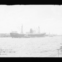 Zwei Frachtschiffe am Hafen mit Kränen, davor ein Schlepper und welliges Wasser im Vordergrund.