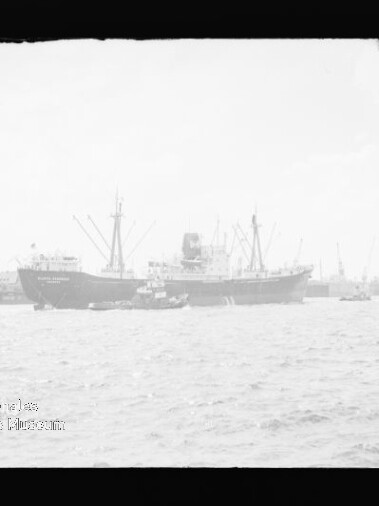 Zwei Frachtschiffe am Hafen mit Kränen, davor ein Schlepper und welliges Wasser im Vordergrund.