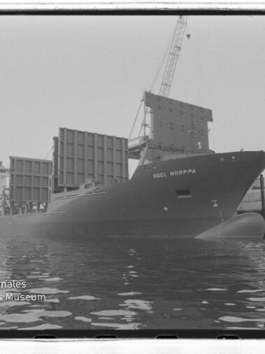 Schwarzweißaufnahme: Containerschiff OOCL NORPPA am Kai, beladene Containerstapel, Hafenkran und Spiegelung im Wasser.