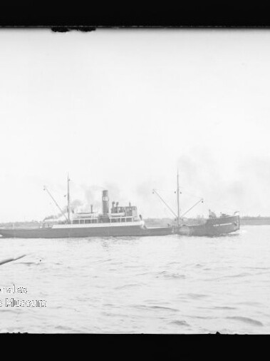 Schwarzweißaufnahme eines Dampfschiffs seitlich auf ruhigem Wasser, rauchender Schornstein und Küstenlinie im Hintergrund.