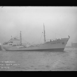 Weißes Frachtschiff mit zwei Masten und Flagge in nebligem Hafen, Wasserzeichen Internationales Maritimes Museum Hamburg.