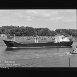 Schwarzweißfoto eines Frachtschiffs auf einem Kanal vor bewaldetem Ufer mit sichtbarem Filmrand und Museumswasserzeichen