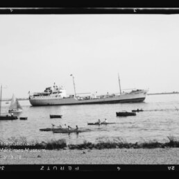 Großes weißes Schiff auf ruhigem Wasser, im Vordergrund mehrere Ruder- und Segelboote und ein Museumstempel.