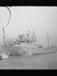 Passagierschiff mit festlicher Beflaggung und Dampf am Bug, begleitet von einem Schlepper im Hafen; Filmnegativ mit Riss.