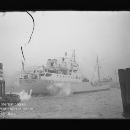 Passagierschiff mit festlicher Beflaggung und Dampf am Bug, begleitet von einem Schlepper im Hafen; Filmnegativ mit Riss.
