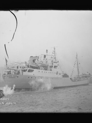 Passagierschiff mit festlicher Beflaggung und Dampf am Bug, begleitet von einem Schlepper im Hafen; Filmnegativ mit Riss.