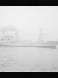 Beflaggtes Frachtschiff mit markantem Mast fährt im nebligen Hafen vor einem großen Hafenkran vorbei.