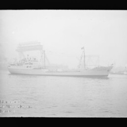 Beflaggtes Frachtschiff mit markantem Mast fährt im nebligen Hafen vor einem großen Hafenkran vorbei.