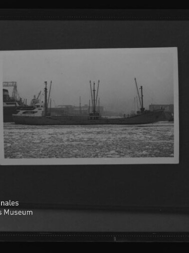 Schwarzweißfoto eines Frachtschiffs mit mehreren Ladekränen im Hafen, im Vordergrund vereistes Wasser und Museumslogo