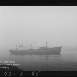 Frachtschiff im dichten Nebel auf ruhigem Wasser, mit Internationales Maritimes Museum Hamburg‑Logo unten links.