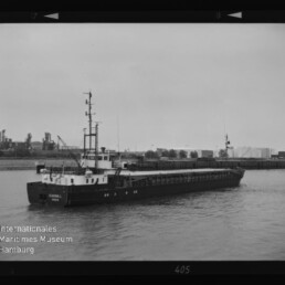 Frachtschiff mit flachem Ladedeck und kleinem Steuerhaus auf ruhigem Hafenwasser, Industriehintergrund, Museumslogo links