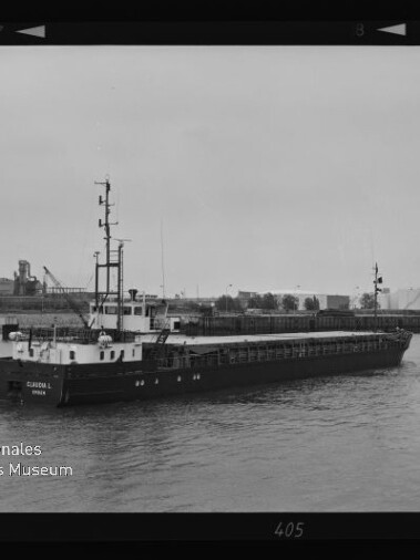 Frachtschiff mit flachem Ladedeck und kleinem Steuerhaus auf ruhigem Hafenwasser, Industriehintergrund, Museumslogo links