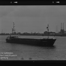 Schwarzweißfoto eines Frachtschiffs vor einem Industriehafen mit Wasserzeichen des Maritimen Museums Hamburg.