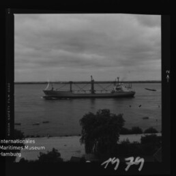 Schwarzweißfoto eines Frachtschiffs auf ruhigem Wasser vor bewölktem Himmel; Ufer mit Bäumen und Museumslogo unten links.