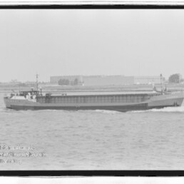 Schwarzweiß-Frachtschiff fährt auf ruhiger See vor flachem Industrie- und Stadthorizont mit ersichtlichem Museumswasserzeichen.