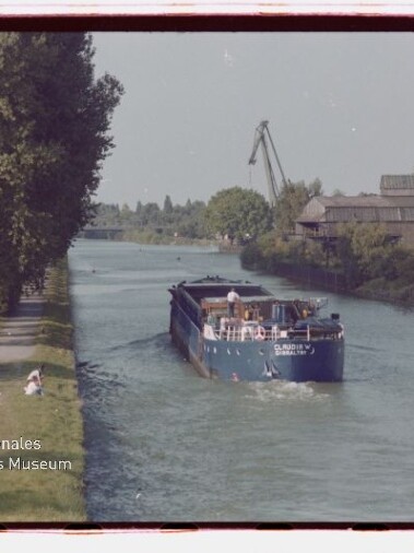 Binnenschiff im Kanal, rechts Industriegebäude, links Baumreihe; unten links Wasserzeichen Internationales Maritimes Museum Hamburg