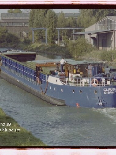 Blaues Binnenschiff CLAUDIA W GIBRALTAR fährt einen Kanal entlang, ein Mann steht auf dem Deck, Industrieanlagen am Ufer.
