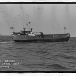 Schwarzweißaufnahme eines Frachters in Seitenansicht auf offener See mit Decksaufbauten und sichtbarem Museumswasserzeichen.