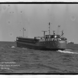 Frachtschiff fährt auf offener See auf die Kamera zu, Bugwelle sichtbar, rechts eine Tonnenboje; Schwarzweißaufnahme mit Museumslogo links unten.