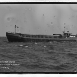 Schwarzweiß-Seitenaufnahme eines Frachtschiffs mit Flaggen auf leicht bewegter See, Museumslogo unten links.