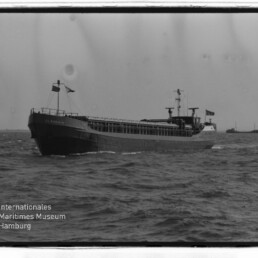 Frachtschiff auf offener See mit weiterem Schiff am Horizont; unten links Logo des Internationalen Maritimen Museums Hamburg.