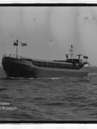 Schwarzweißfoto eines Frachtschiffs auf See mit Flaggen und offenem Laderaum; unten links Museumswasserzeichen.