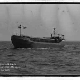 Schwarzweißfoto eines Frachtschiffs auf See mit Flaggen und offenem Laderaum; unten links Museumswasserzeichen.