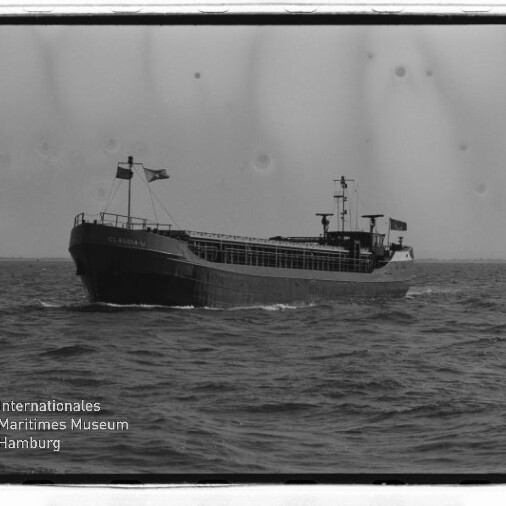 Schwarzweißfoto eines Frachtschiffs auf See mit Flaggen und offenem Laderaum; unten links Museumswasserzeichen.