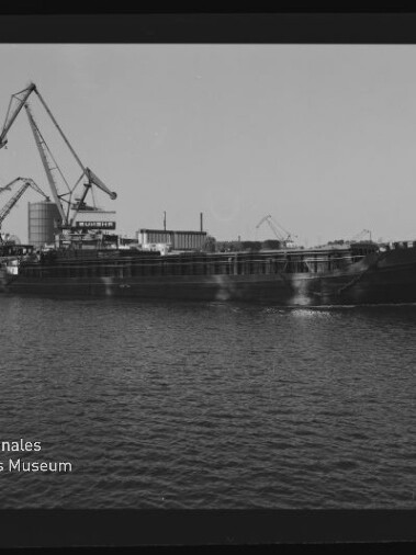 Frachtschiff auf ruhigem Wasser vor einem Industriehafen mit mehreren Hafenkränen; schwarz-weißes Bild mit Museumswasserzeichen