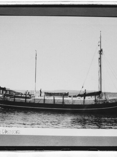 Schwarzweißfoto eines historischen Segelschiffs mit zwei Masten, ruhend vor einer flachen Küstenlinie.