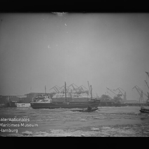 Frachtschiff im Hafen mit Treibeis im Wasser und Kränen im Hintergrund, Museum-Wasserzeichen unten links.
