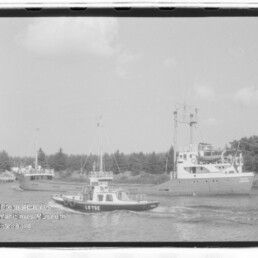 Lotsenboot mit Aufschrift LOTSE fährt vor einem größeren Schiff entlang eines baumbewachsenen Ufers.